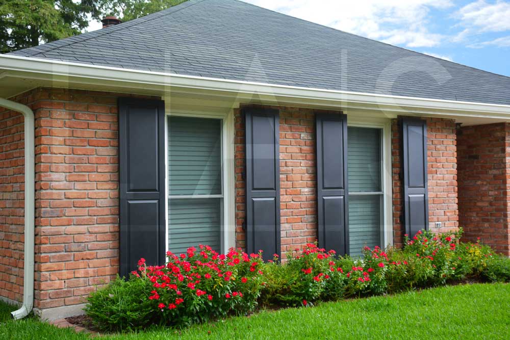 Brick home with raised panel shutters in black, featuring a well-maintained lawn and vibrant flower bed.
